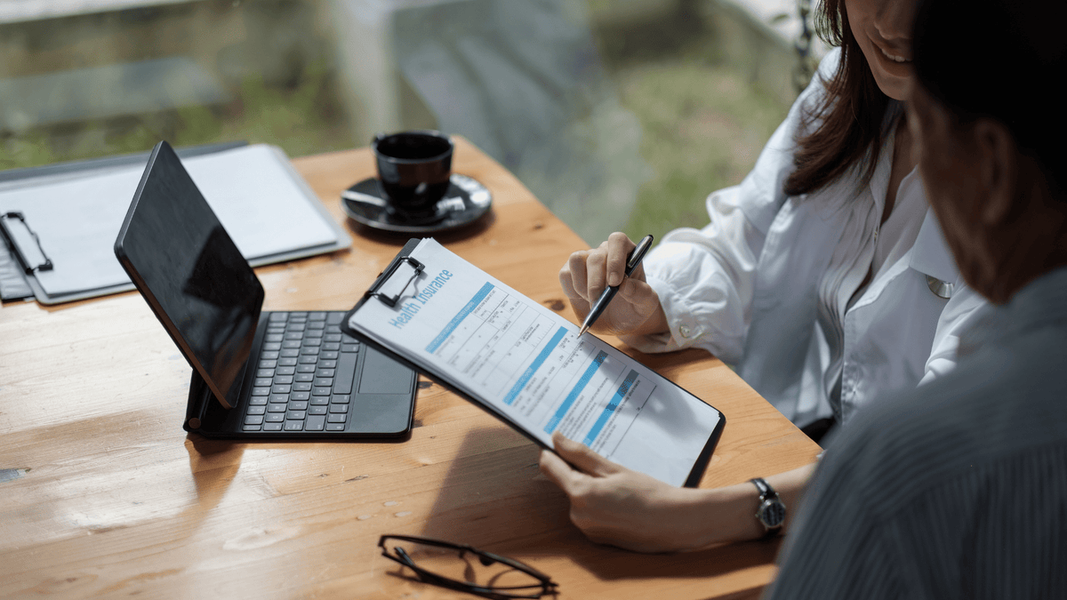 Eine Person, die mit einem Arzt oder Berater über eine Gesundheitsversicherung spricht und ein Formular mit der Aufschrift "Health Insurance" ausfüllt. Auf dem Tisch stehen ein Laptop und eine Kaffeetasse.