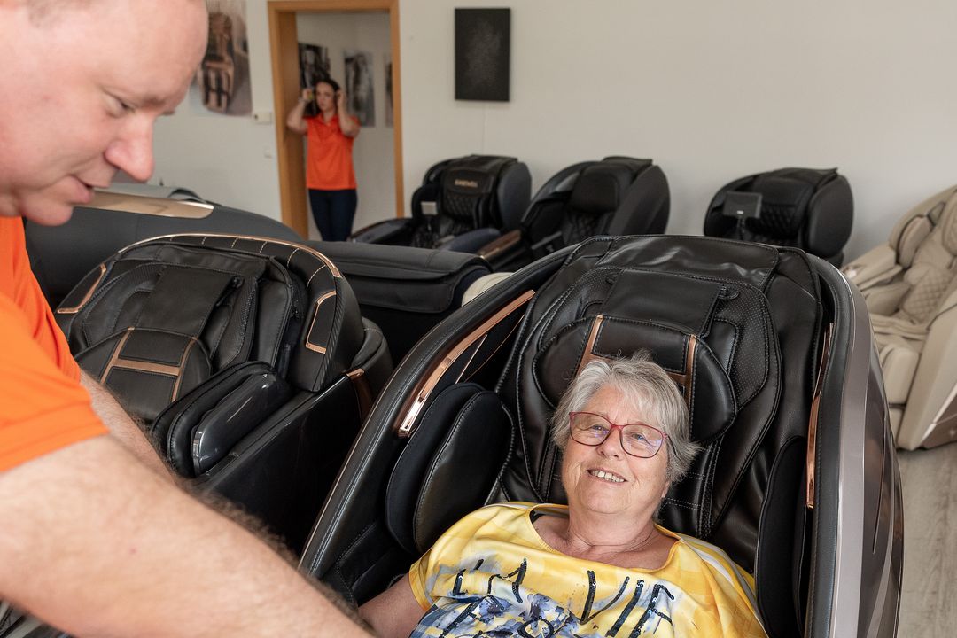 Een adviseur van Massagesessel Welt buigt zich voorover naar een klant die in de massagestoel zit en legt haar uit hoe de stoel werkt