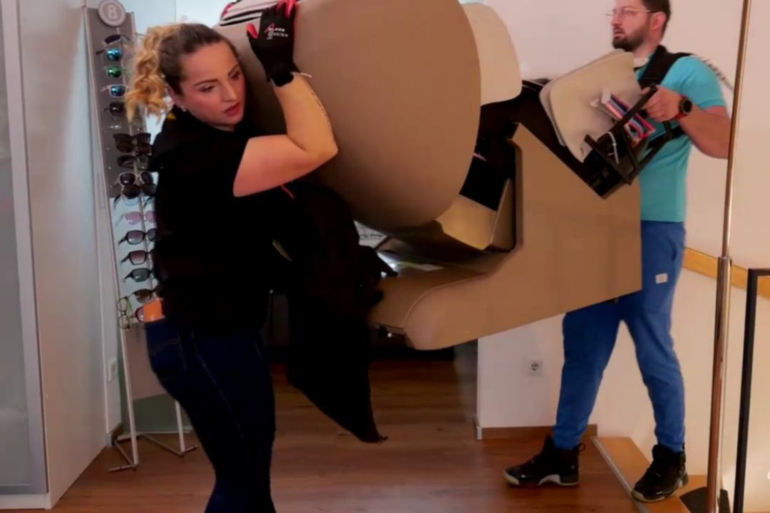 Twee medewerkers van Massagesessel Welt dragen een massagestoel met draagriemen de ruimte binnen waar hij moet komen te staan